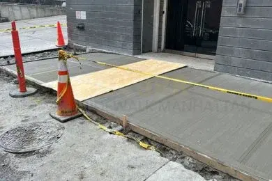 Freshly poured sidewalk slab taped off with traffic cones and a plywood walkway at a building entrance
