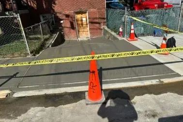 Freshly poured sidewalk in front of a brick house, protected by caution tape and a traffic cone