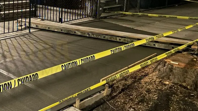 Freshly poured sidewalk at night, framed with wooden forms and caution tape beside an iron fence
